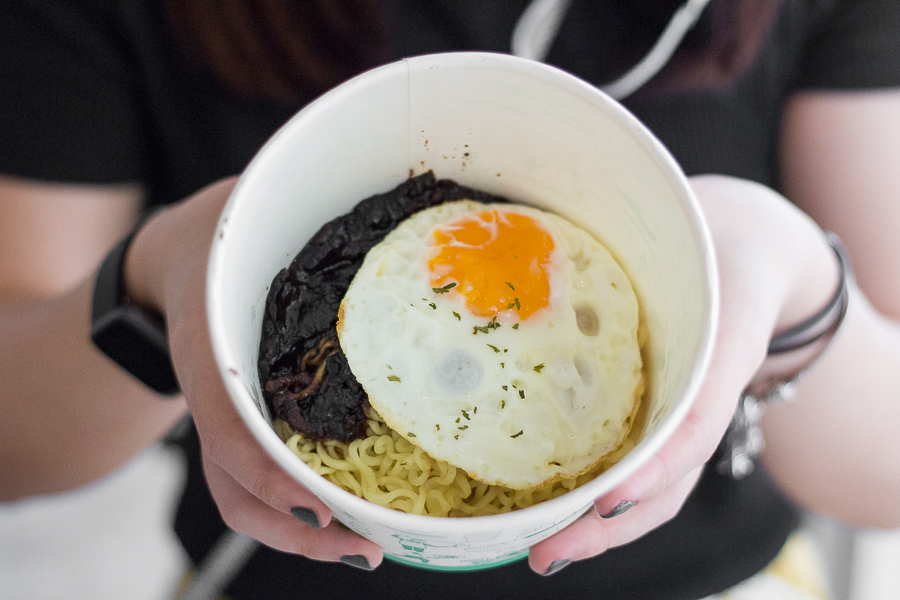 A paper bowl with noodles, a sunny side up and some jjajang sauce