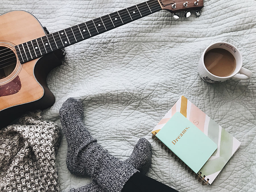 a person on bed with a guitar, a notebook and a cup of coffee