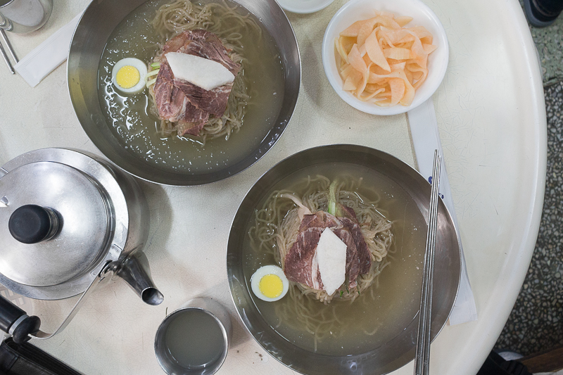 Two bowls of Naengmyeon on a table in a Seoul restaurant with side dishes and a kettle of warm soup