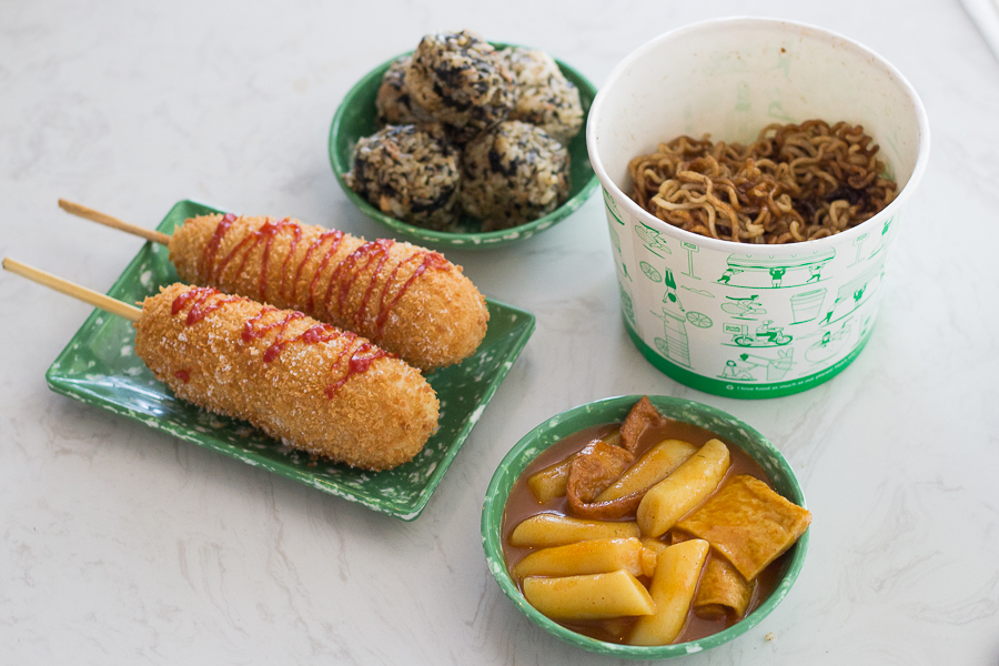 a table of Korean food such as tteokbokki, hot dogs, rice balls and ramyeon