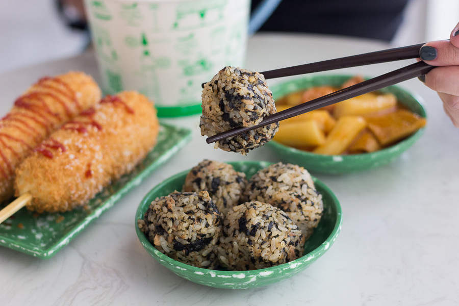 Chopsticks holding up a tuna mayo rice ball with hotdogs and tteokbokki in the background