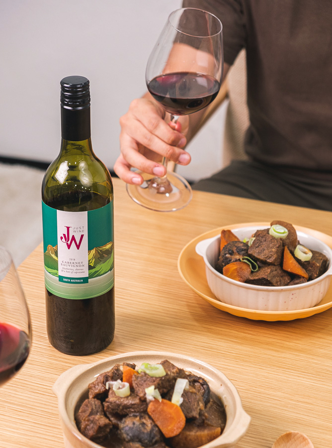 A person holding a glass of Just Wine's Cabernet Sauvignon with bowls of Galbi-jjim