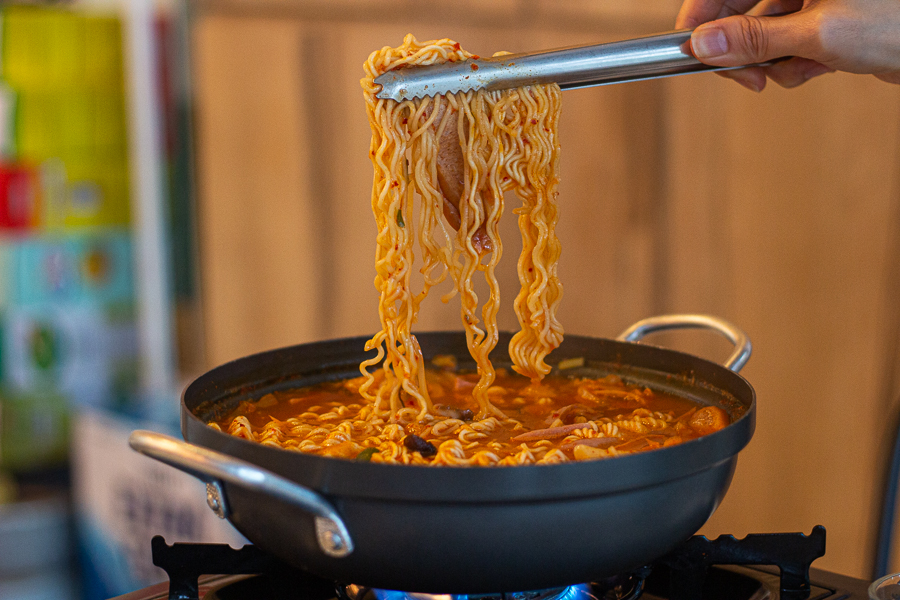 Using a pair of tongs to pick up instant noodles from a bowl of Army Stew