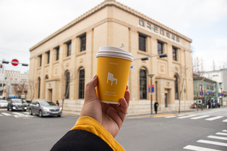 A yellow cup from Nontable with a background of the Daegu Modern History Museum