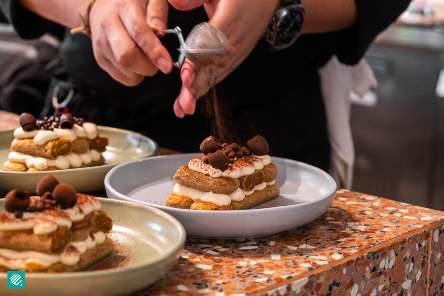 A person sprinkling cocoa powder over a cold drip tiramisu